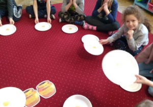 Dzieci siedzą na dywanie. Pokazują talerzyki z miodem